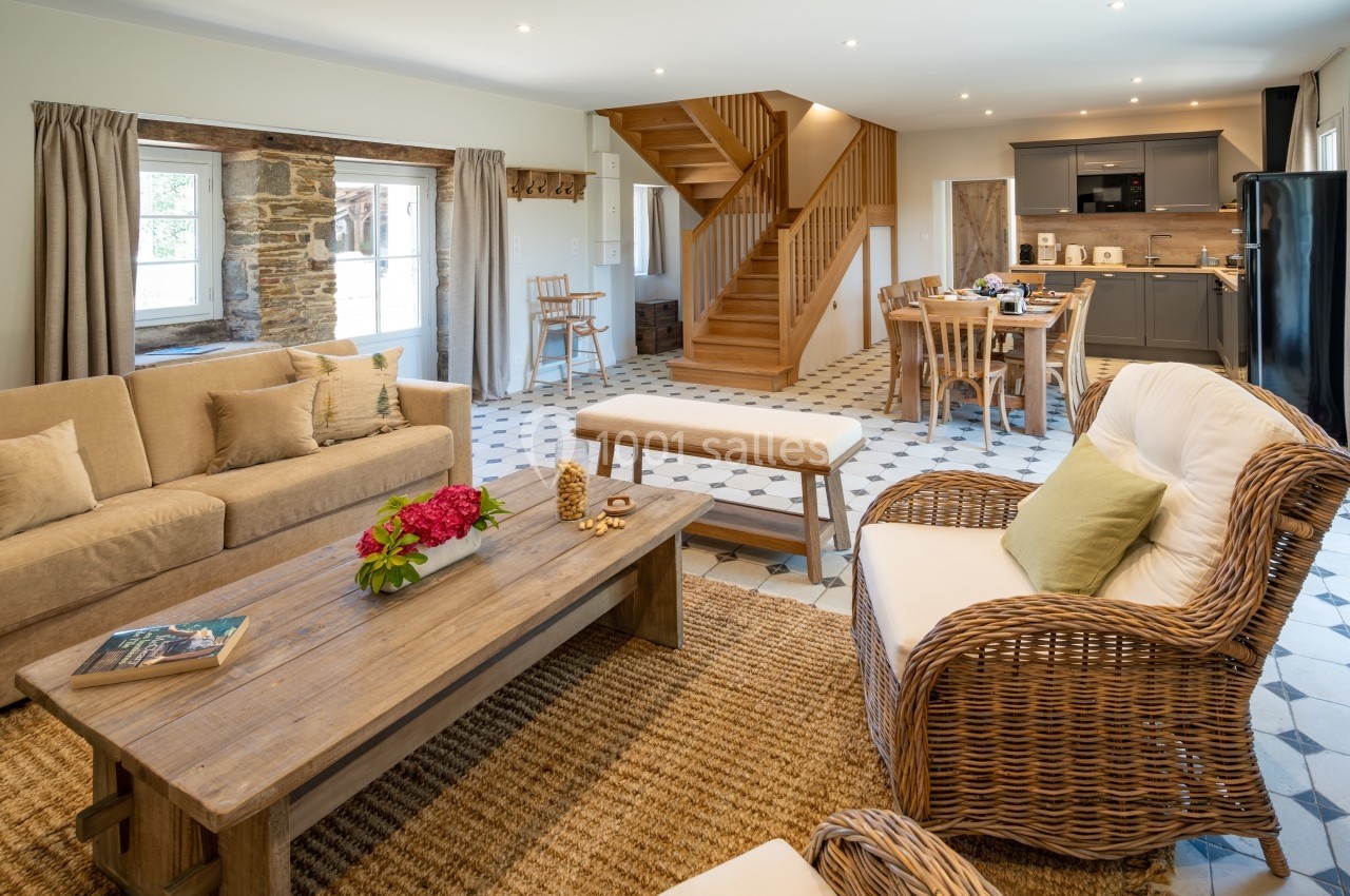 Salon lumineux avec canapé beige, fauteuils en rotin, table basse en bois, cuisine ouverte et escalier en bois.