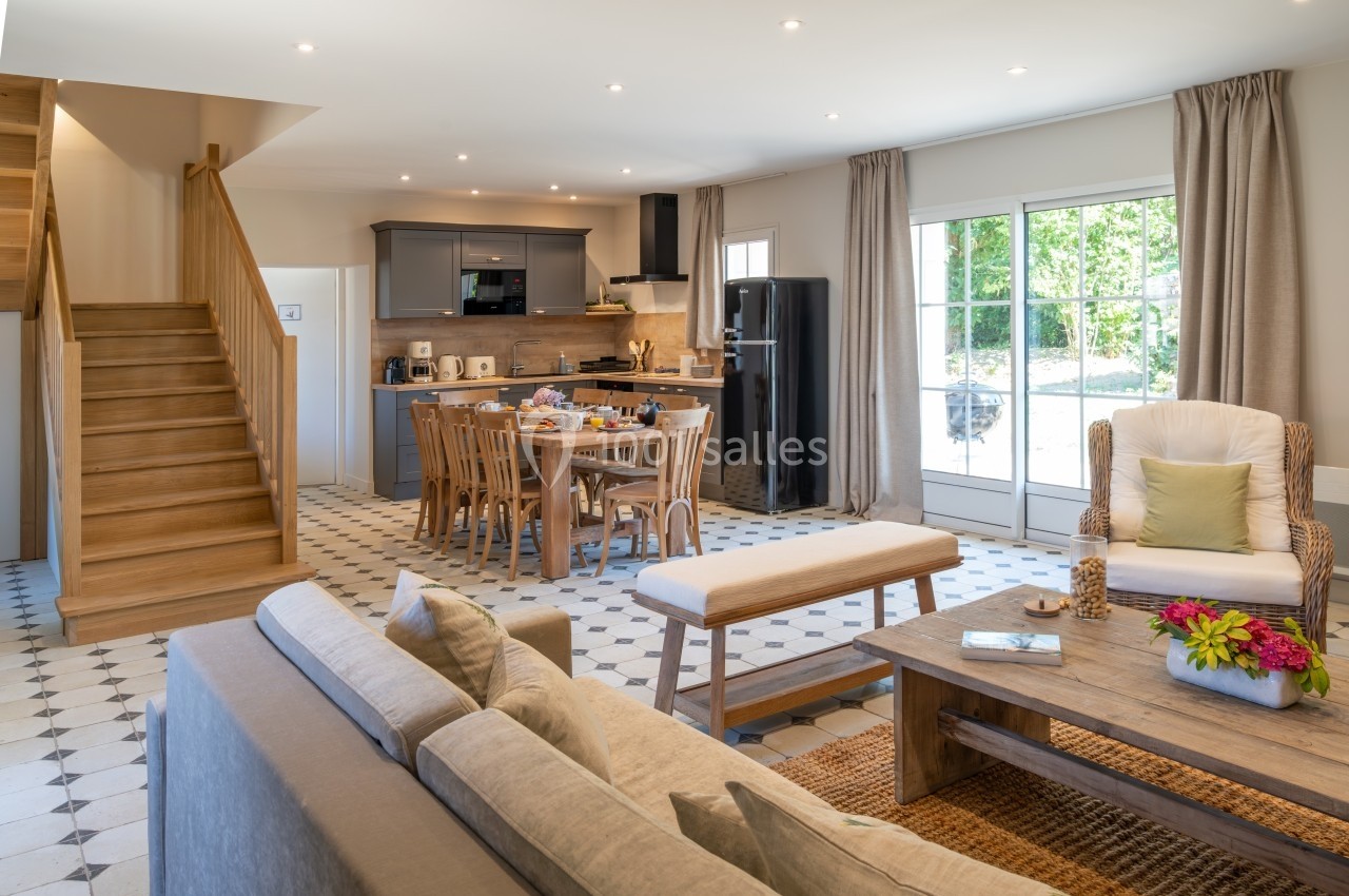 Salon lumineux avec cuisine ouverte, table en bois, canapé beige, escalier en bois et grandes fenêtres donnant sur un jardin.