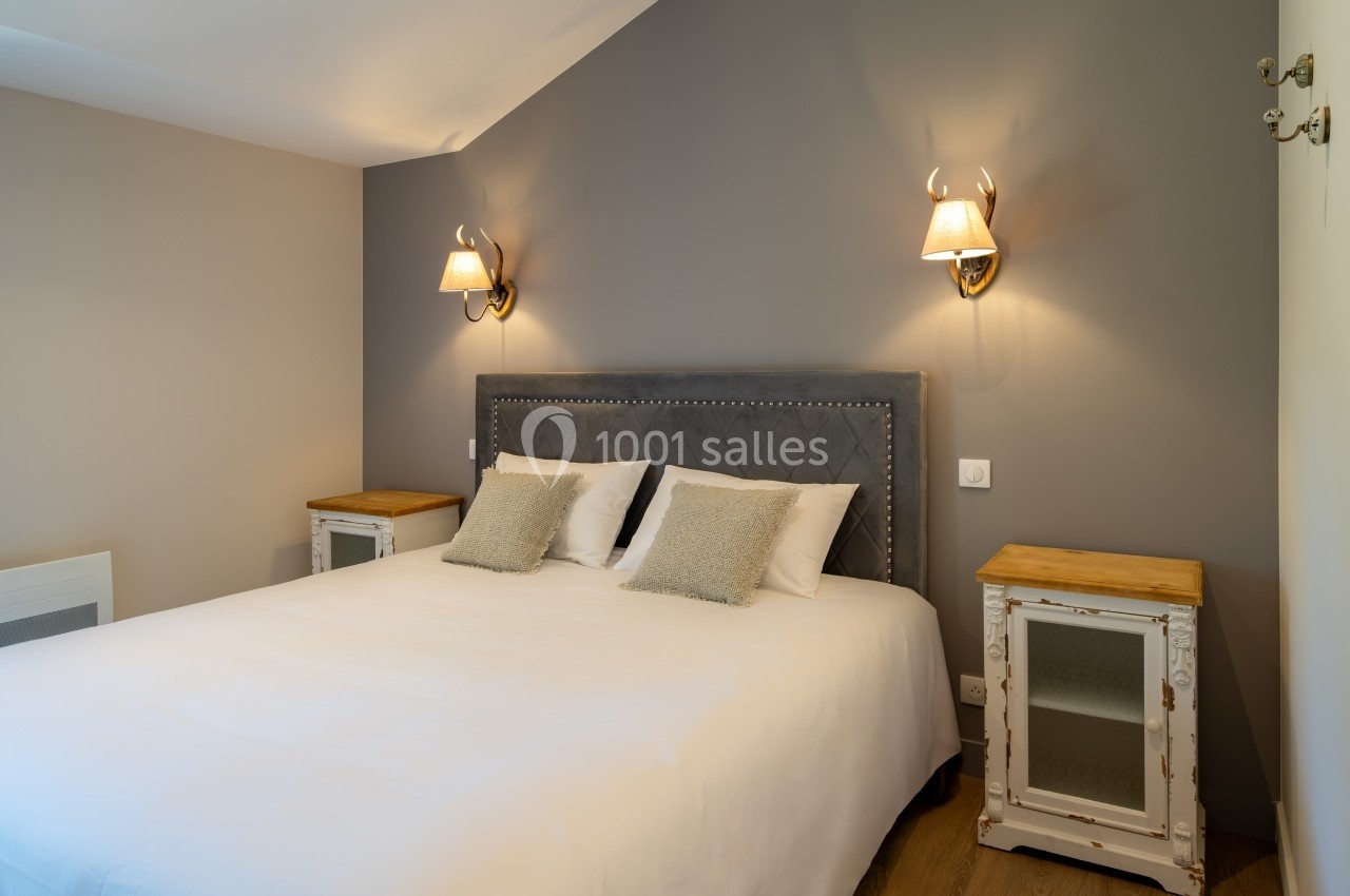 Chambre avec lit double, tête de lit capitonnée, tables de chevet en bois et appliques murales sur un mur gris.