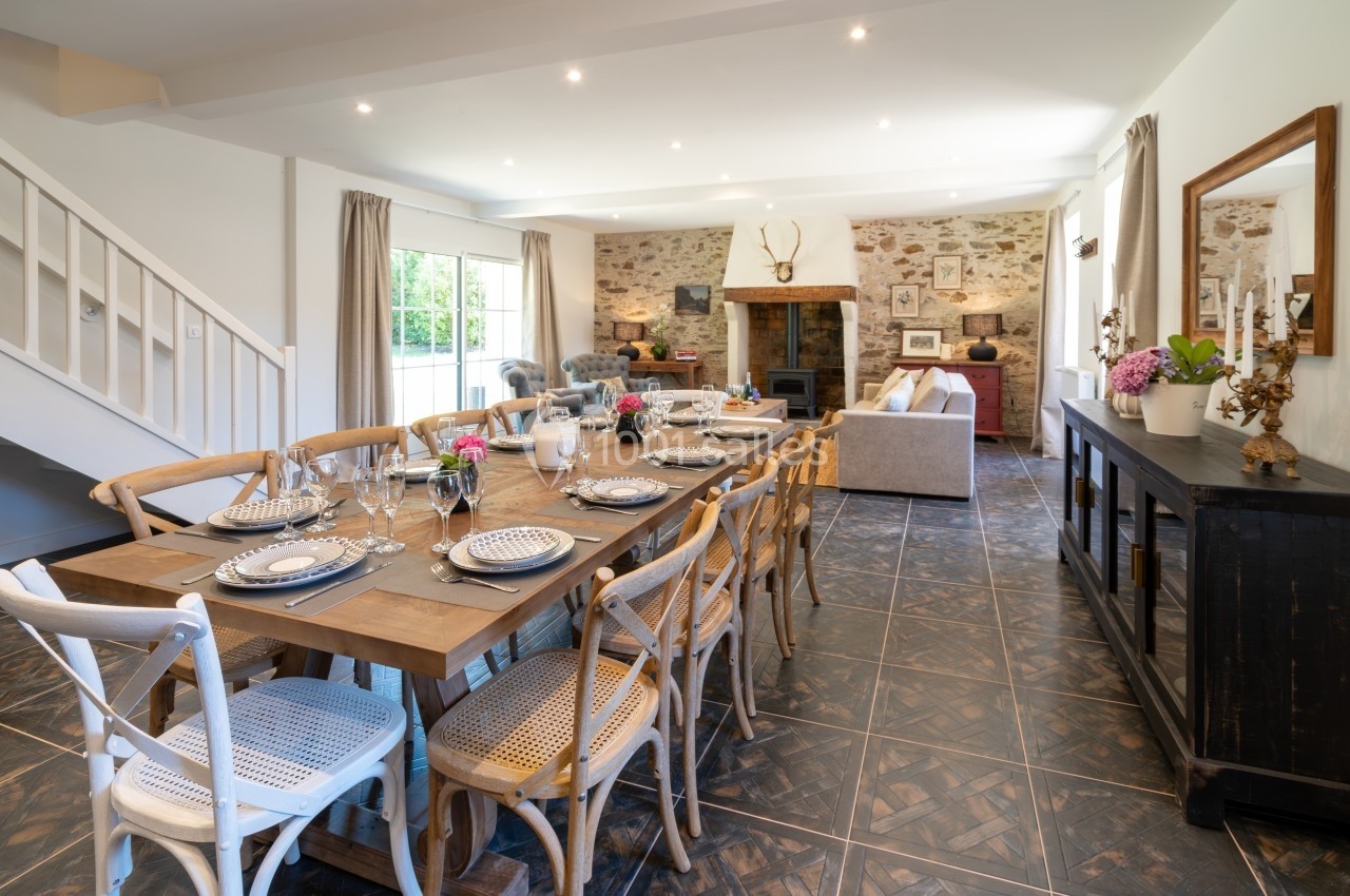 Salle à manger lumineuse avec grande table en bois, chaises en osier, et salon en arrière-plan avec murs en pierre.