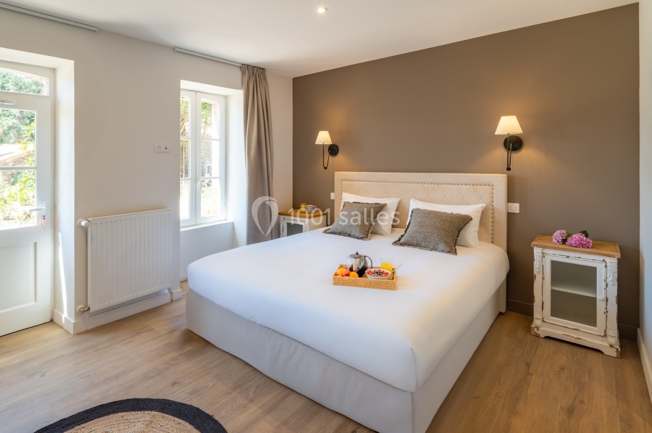 Chambre lumineuse avec lit double, murs beige et taupe, tables de chevet, et plateau de fruits sur le lit.