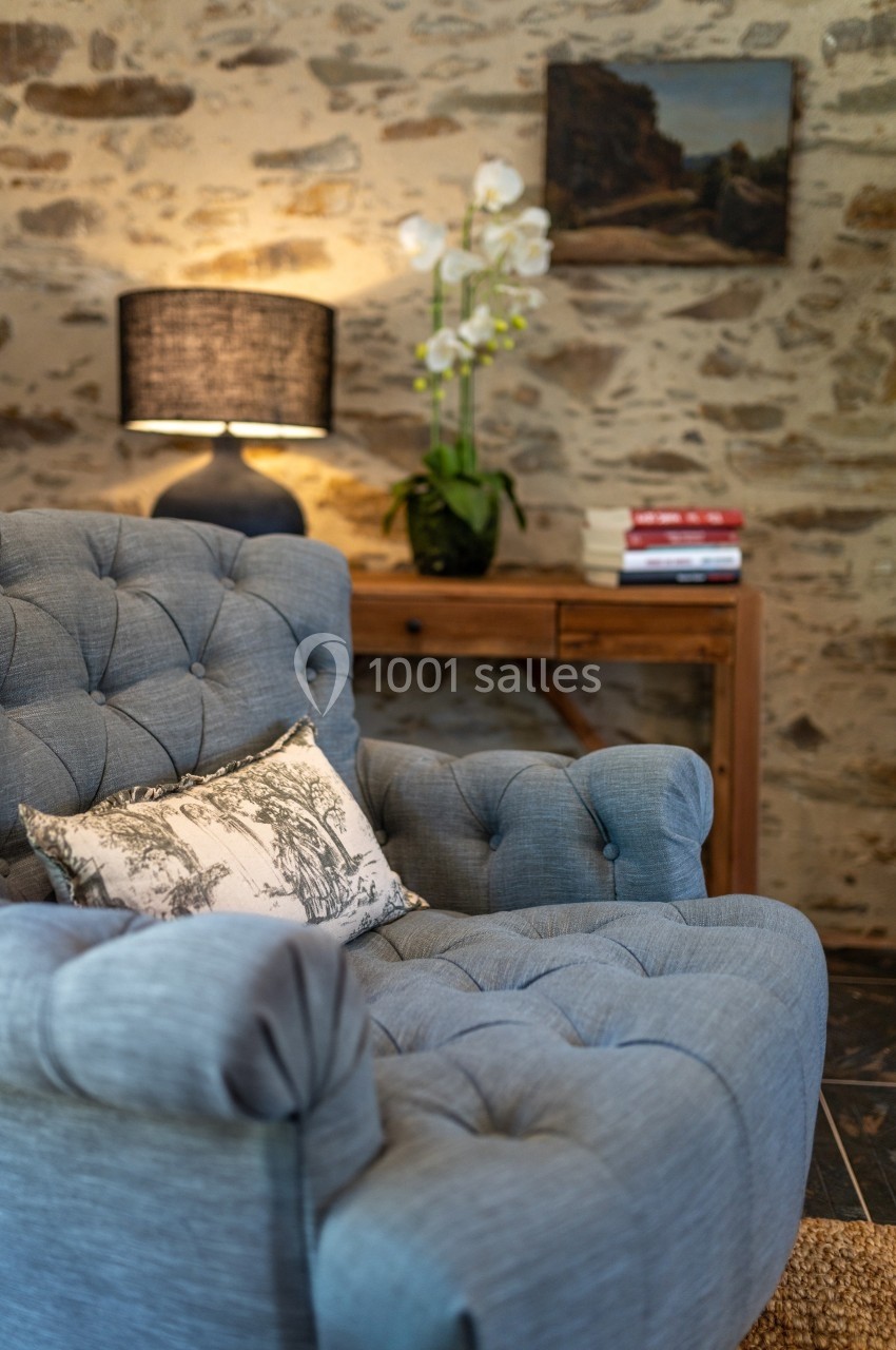 Fauteuil bleu capitonné devant un mur en pierre, avec une lampe, une orchidée et des livres sur une table en bois.