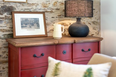 Commode rouge surmontée de cadres, lampe noire et carafe, avec un mur en pierre en arrière-plan.