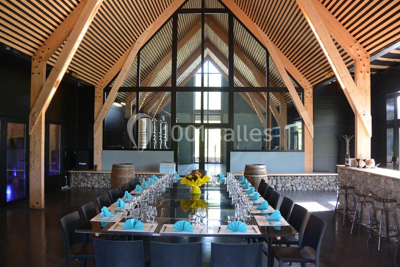 Salle de réception moderne avec charpente en bois apparente, grande table dressée et baies vitrées en arrière-plan.