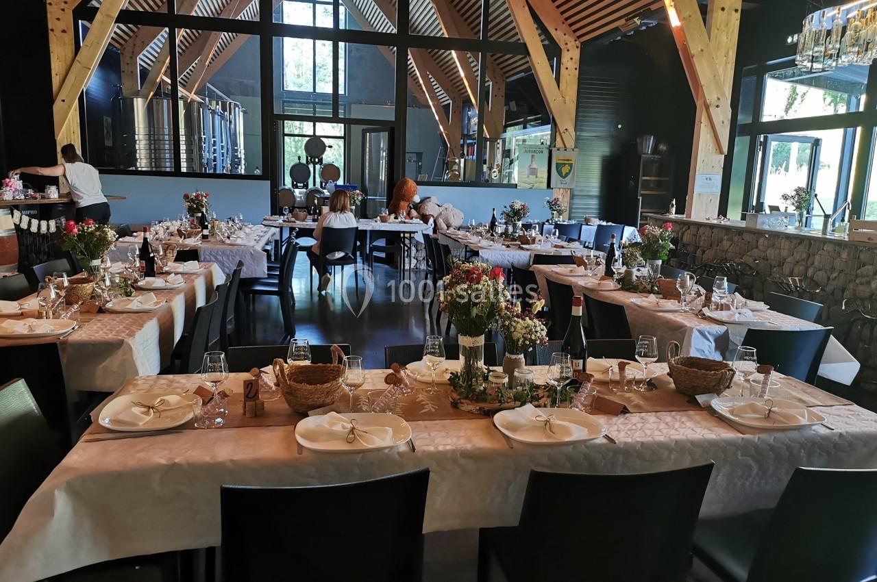 Salle de réception avec tables dressées, décorées de fleurs et de paniers, dans un espace lumineux avec charpente en bois.