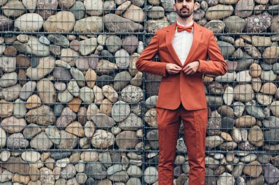 Une femme en robe de mariée sourit face à un homme en costume orange dans un vignoble verdoyant.