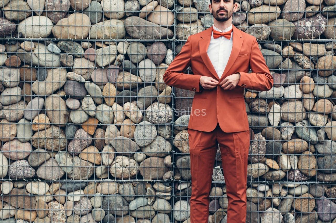 Homme en costume orange ajustant sa veste devant un mur de pierres grillagées.