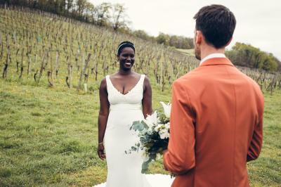 Une femme en robe de mariée sourit face à un homme en costume orange dans un vignoble verdoyant.