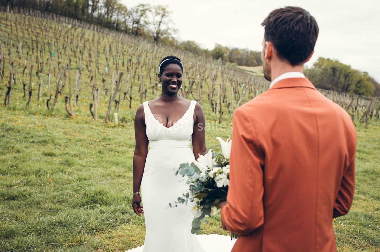 Une femme en robe de mariée sourit face à un homme en costume orange dans un vignoble verdoyant.