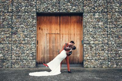 Une femme en robe de mariée sourit face à un homme en costume orange dans un vignoble verdoyant.
