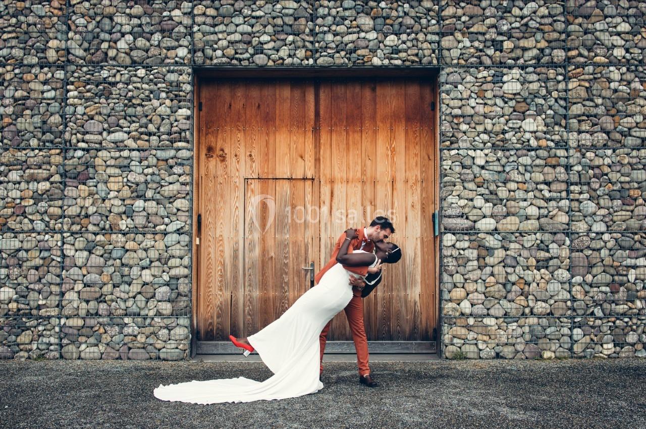 Un couple en tenue de mariage s'embrasse devant une porte en bois encadrée par un mur de pierres.