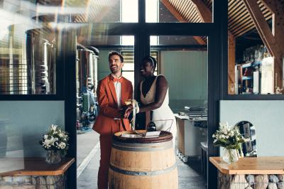 Une femme en robe de mariée sourit face à un homme en costume orange dans un vignoble verdoyant.