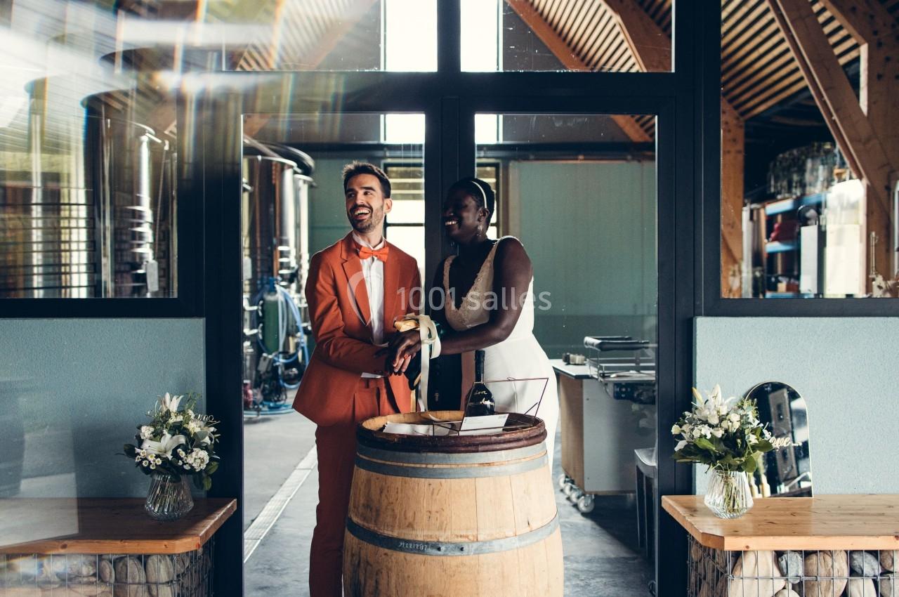 Un couple habillé élégamment, debout près d'un tonneau en bois, dans un espace lumineux avec des fleurs et des équipements…