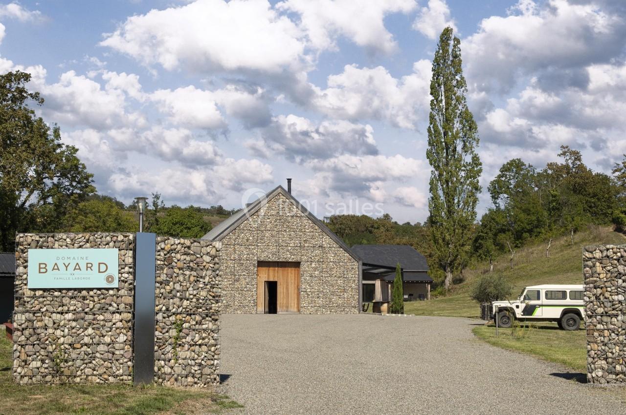 Bâtiment en pierre entouré de verdure avec une entrée gravillonnée et un panneau indiquant ’Domaine Bayard’.