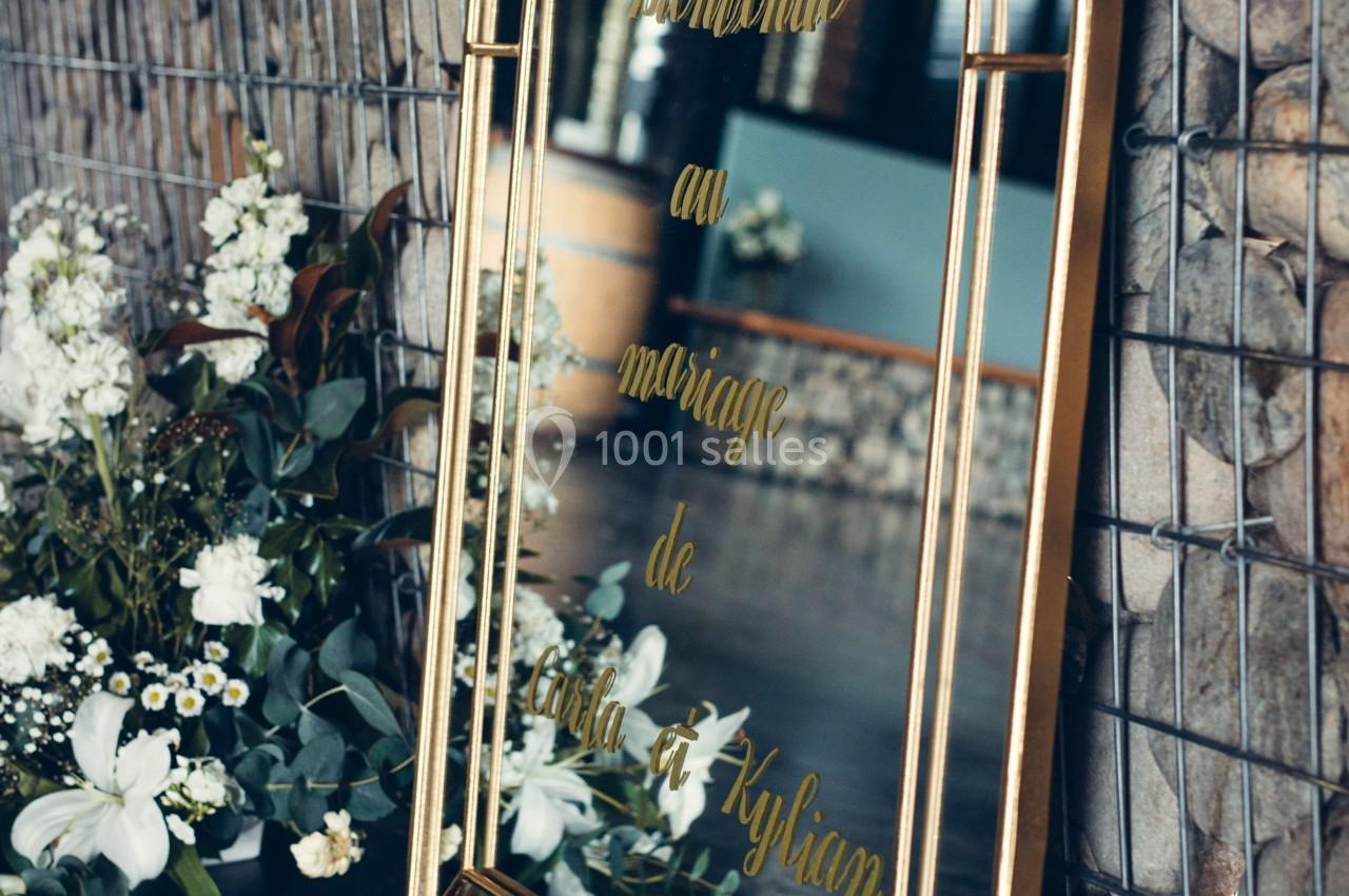 Miroir doré avec inscription ’Bienvenue au mariage de Julia et Kylian’, entouré de fleurs blanches et feuillage.
