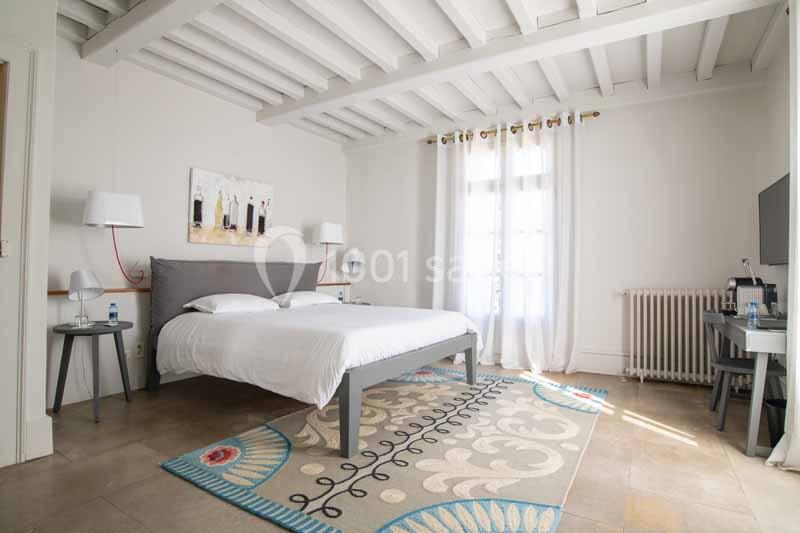 Chambre lumineuse avec lit double, tapis coloré, bureau, lampes et plafond en poutres apparentes blanches.