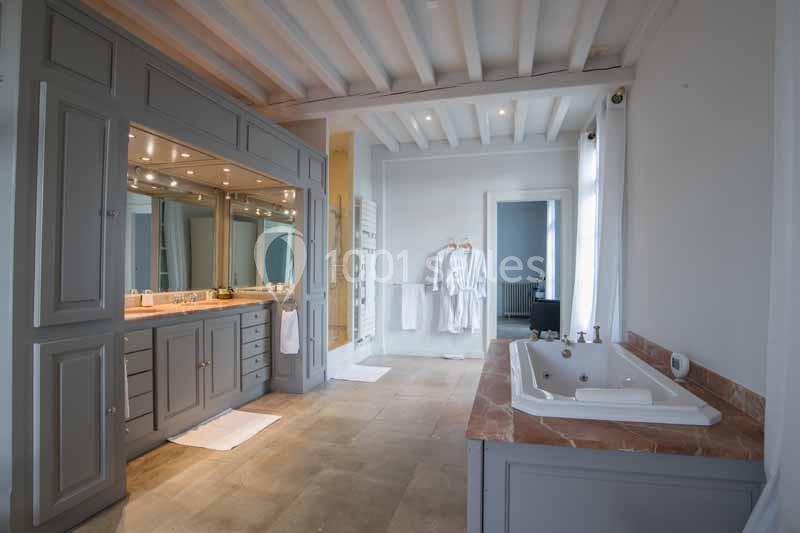 Salle de bain spacieuse avec baignoire encastrée, meuble gris à miroirs et peignoirs suspendus près d'une fenêtre.