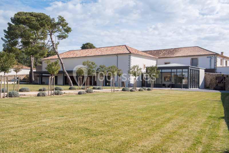 Bâtiment rénové avec façade blanche, toit en tuiles, grande verrière et jardin avec pelouse et jeunes arbres.