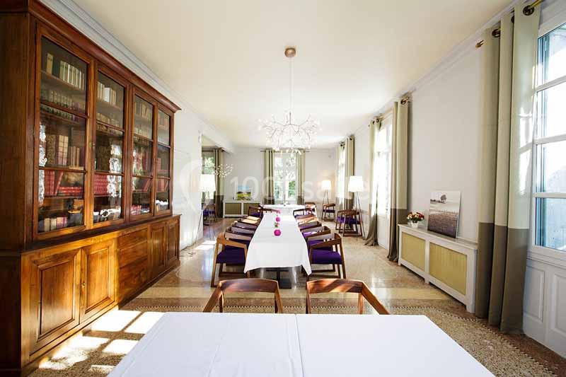 Salle lumineuse avec tables dressées, chaises en bois, grande bibliothèque vitrée et fenêtres laissant entrer la lumière…
