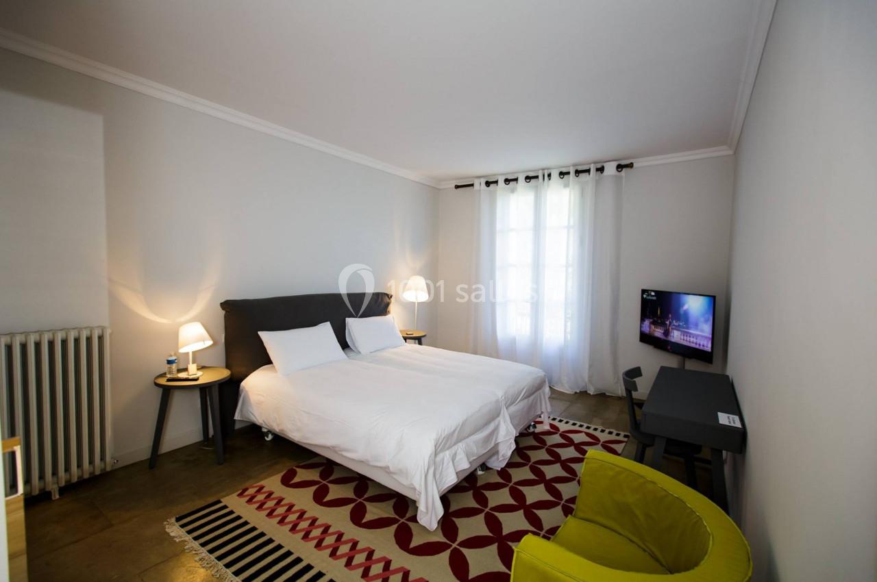 Chambre lumineuse avec lit double, bureau, fauteuil vert, tapis à motifs et fenêtre avec rideaux blancs.