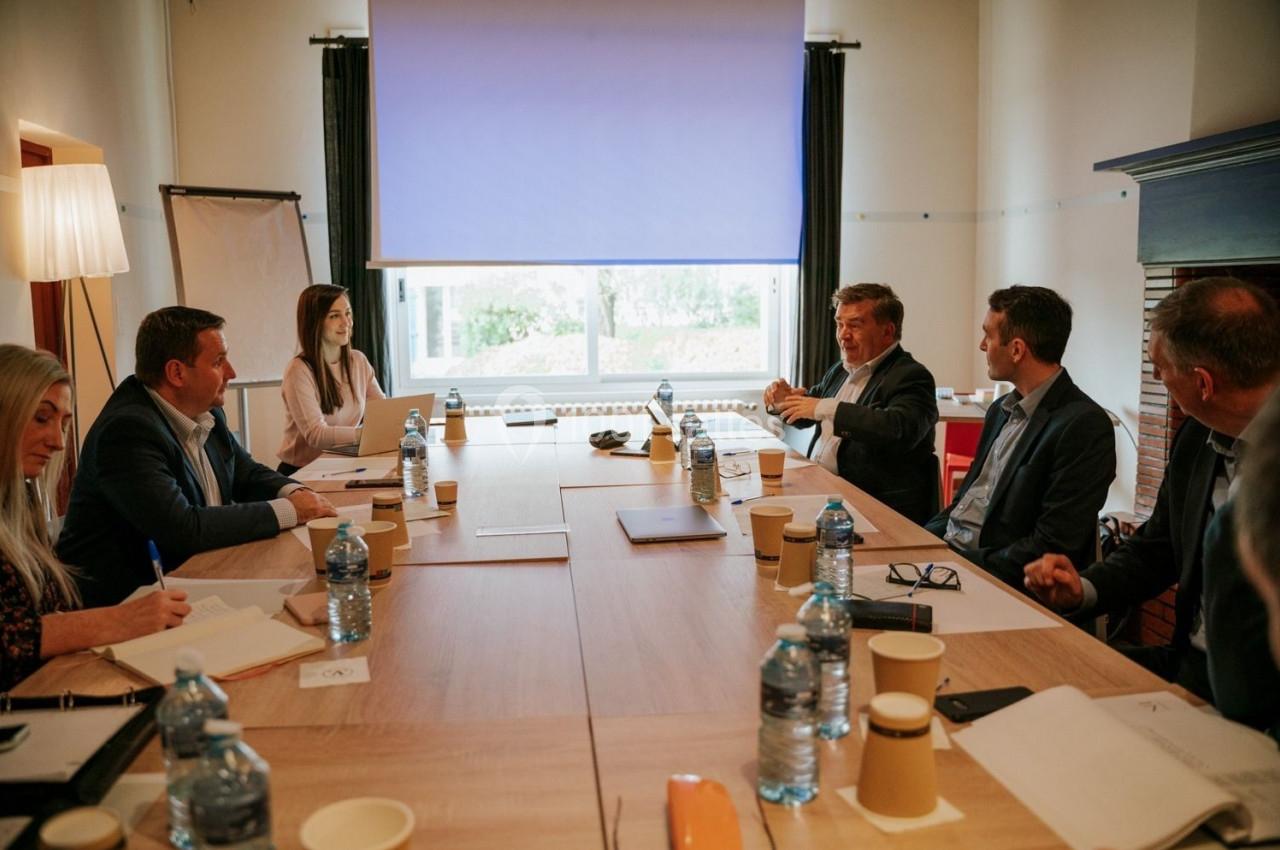 Réunion d'affaires autour d'une grande table, plusieurs personnes discutent dans une salle lumineuse avec un écran.