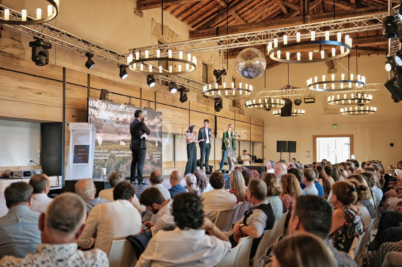 Des intervenants debout sur une scène face à un public dans une salle avec des lustres et une charpente en bois apparente.
