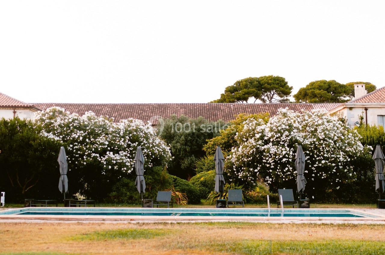 Vue d'une grande maison entourée de végétation, avec une piscine et des chaises longues au premier plan.