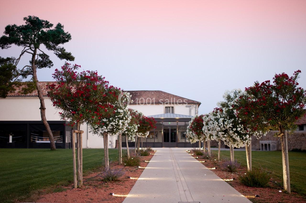 Allée bordée d'arbres fleuris menant à un bâtiment avec toit en tuiles sous un ciel rose au crépuscule.