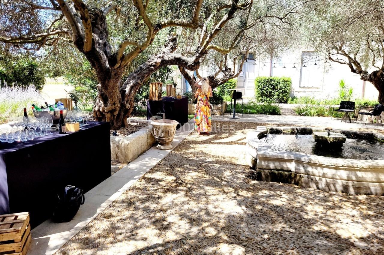 Jardin ombragé avec fontaine centrale, tables noires dressées pour un événement et une personne en robe colorée.