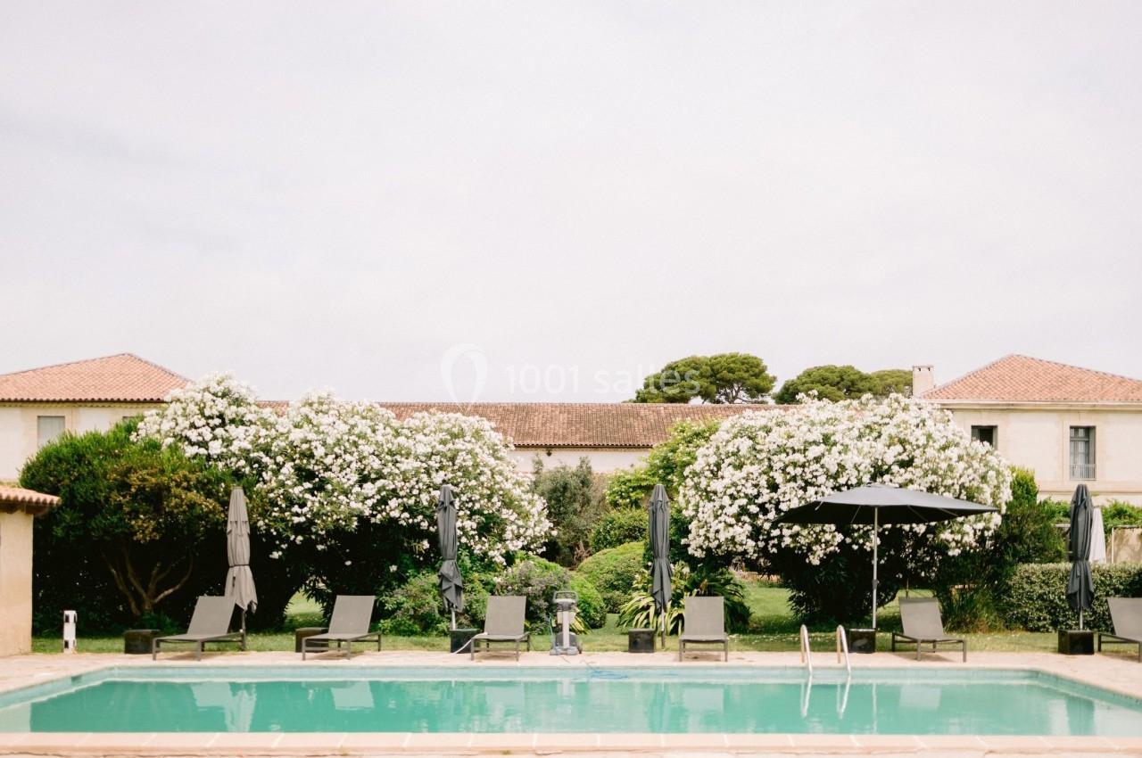 Piscine extérieure entourée de transats, parasols noirs et arbustes fleuris, avec des bâtiments en arrière-plan.
