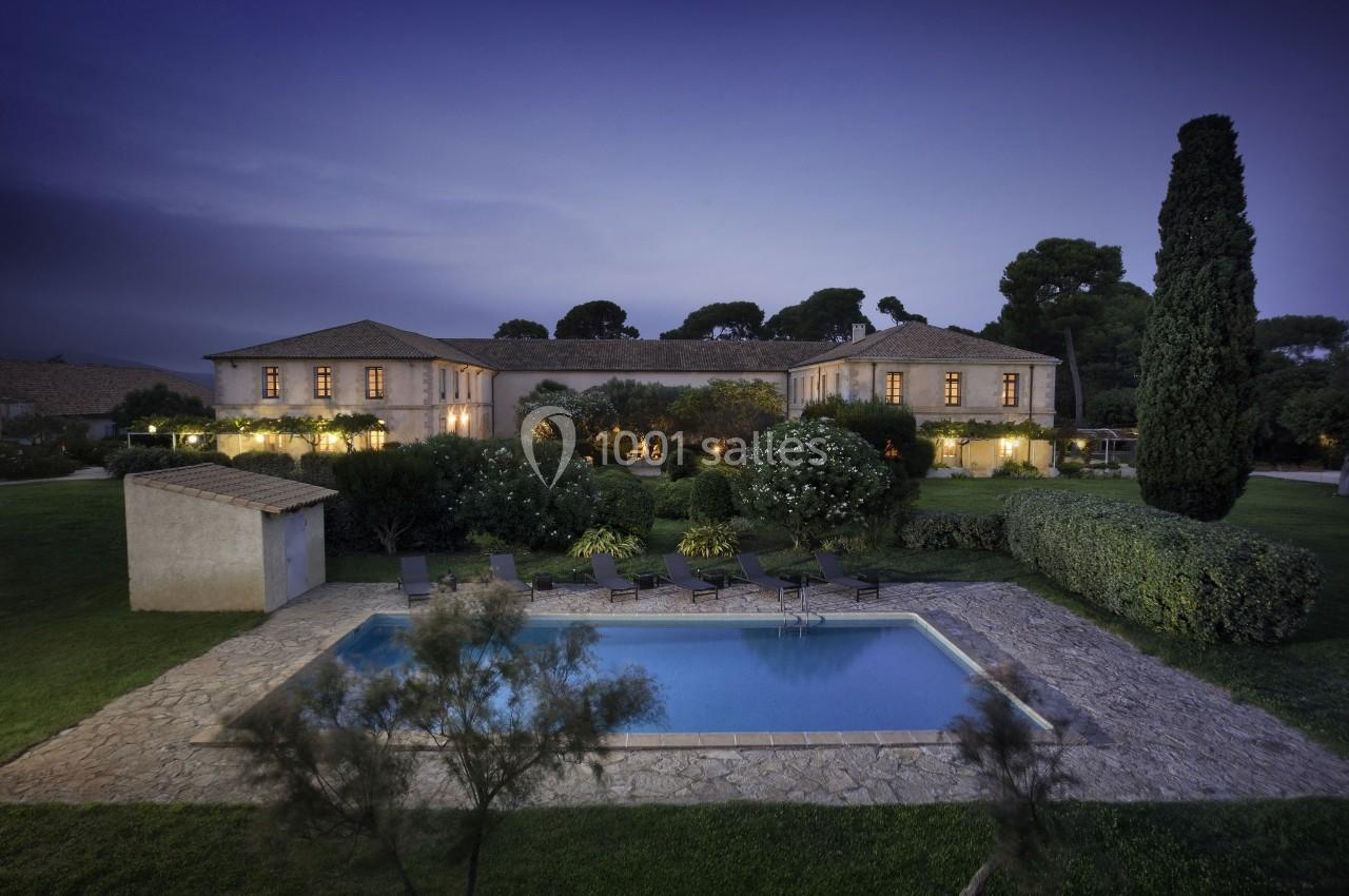 Vue d'une grande maison en pierre entourée de jardins, avec une piscine éclairée au premier plan, au crépuscule.