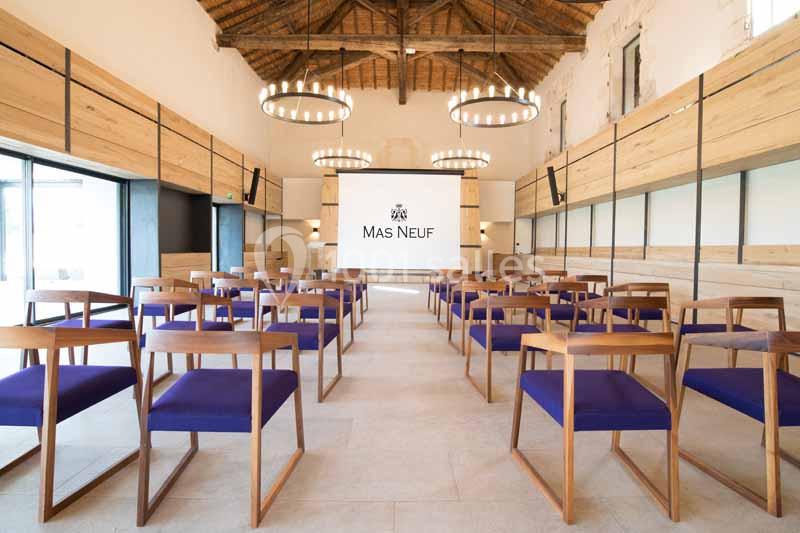 Salle de conférence lumineuse avec des chaises en bois et tissu bleu, écran de projection et poutres apparentes au plafond.