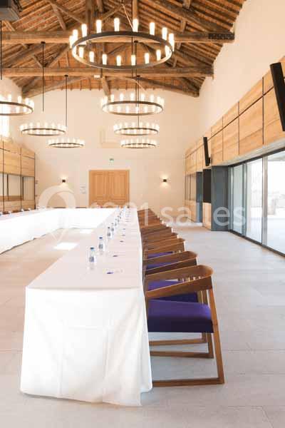 Salle de réunion lumineuse avec une longue table blanche, des chaises en bois et un plafond en poutres apparentes.