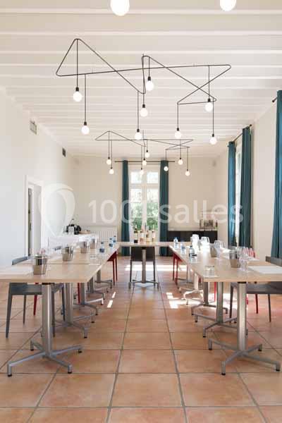 Salle lumineuse avec tables disposées en U, chaises colorées, grandes fenêtres et luminaires suspendus modernes.