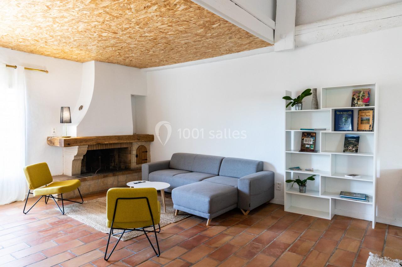 Salon lumineux avec canapé gris, fauteuils jaunes, bibliothèque blanche, cheminée rustique et sol en carrelage.