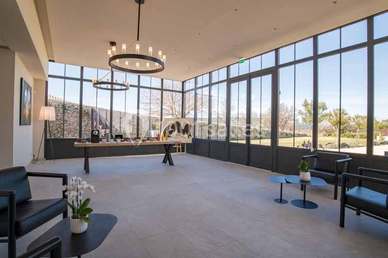 Salle lumineuse avec de grandes baies vitrées, mobilier moderne, tables décorées et vue sur un extérieur arboré.