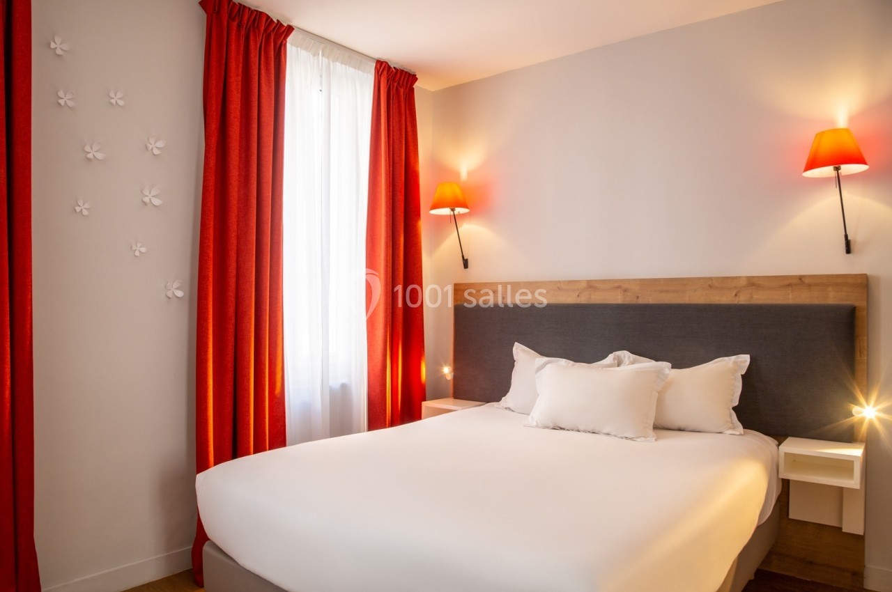 Chambre avec lit double, tête de lit en bois, murs clairs, rideaux rouges et appliques murales avec lumière chaude.