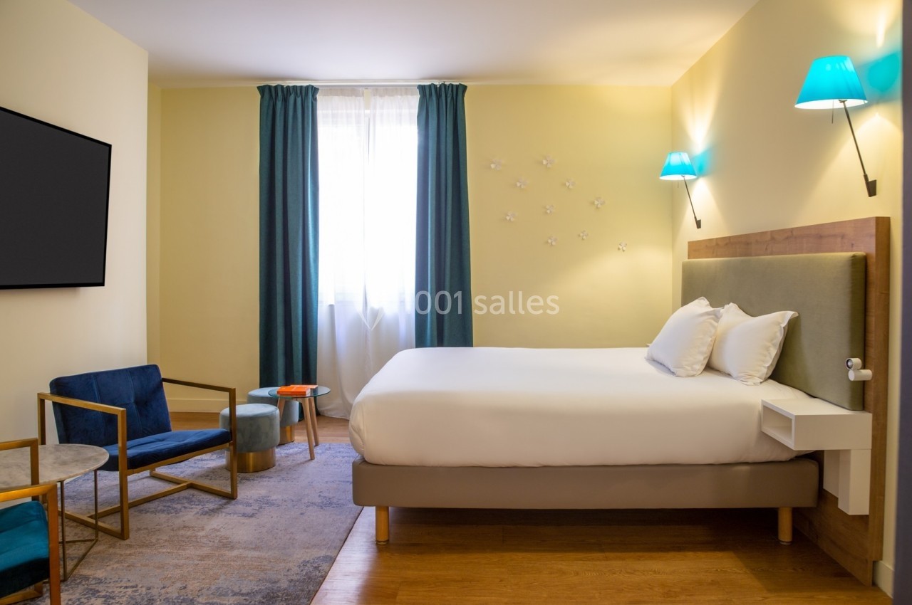 Chambre lumineuse avec lit double, fauteuils, table basse, rideaux bleus et appliques murales dans un décor moderne.