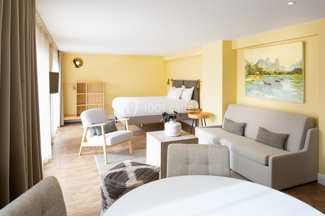 Chambre lumineuse avec lit double, canapé, fauteuils, table ronde et décoration murale dans un intérieur aux tons jaunes.