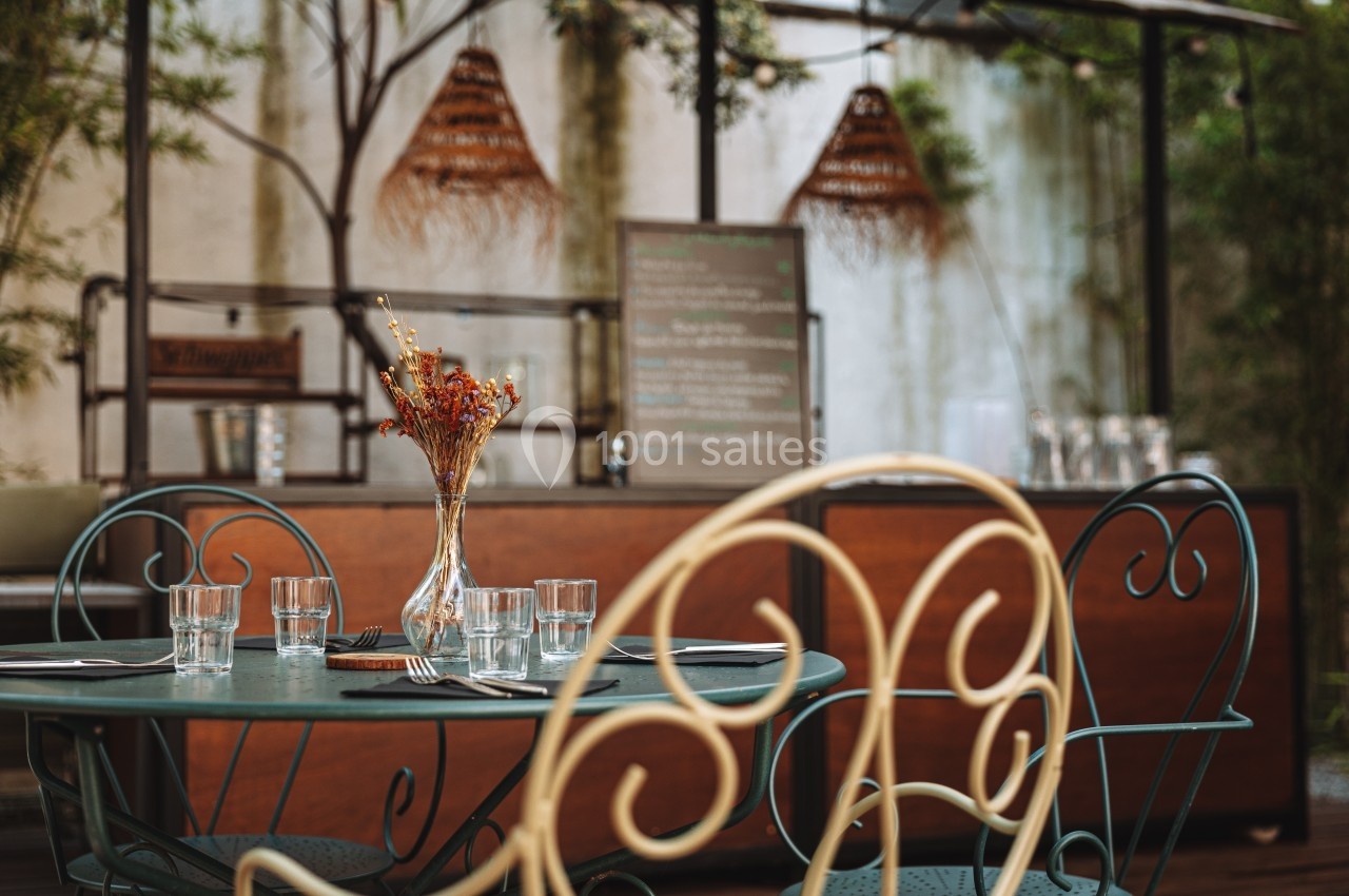 Table en métal avec verres et couverts, décorée d'un vase de fleurs séchées, dans une terrasse extérieure chaleureuse.