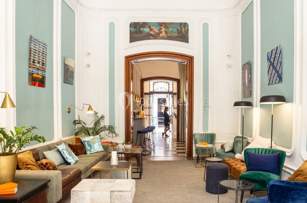 Salon lumineux avec canapés, fauteuils colorés, tables basses et vue sur un couloir menant à une entrée vitrée.