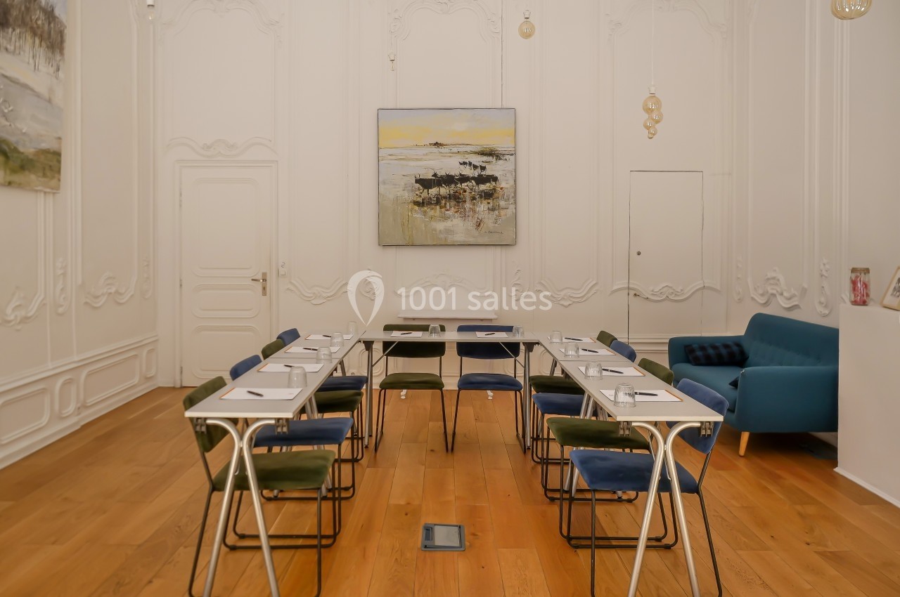 Salle de réunion lumineuse avec tables disposées en U, chaises colorées, parquet en bois et tableaux accrochés aux murs.