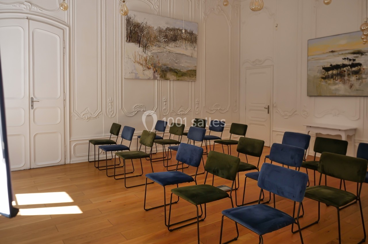 Salle lumineuse avec parquet, chaises alignées bleu et vert, murs ornés de moulures et tableaux paysagers.