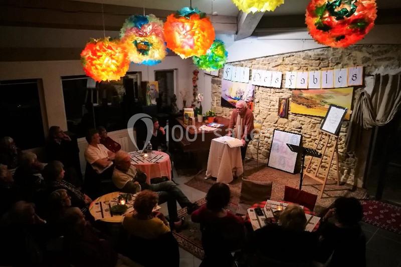 Un conteur s'adresse à un public assis dans une salle décorée de lampions colorés et d'affiches.