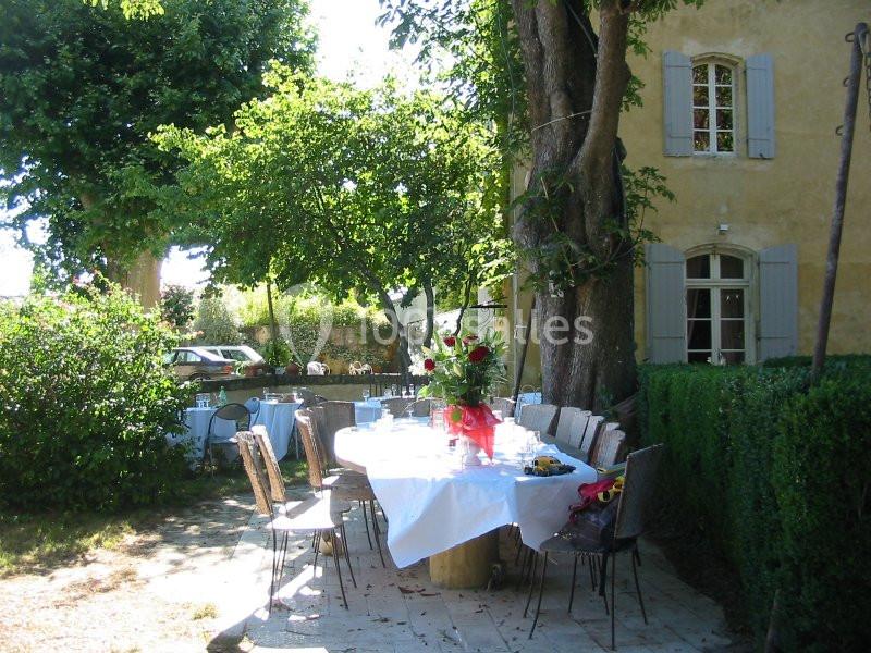 Table dressée avec nappe blanche et bouquet de fleurs rouges dans un jardin ombragé près d'une maison en pierre.