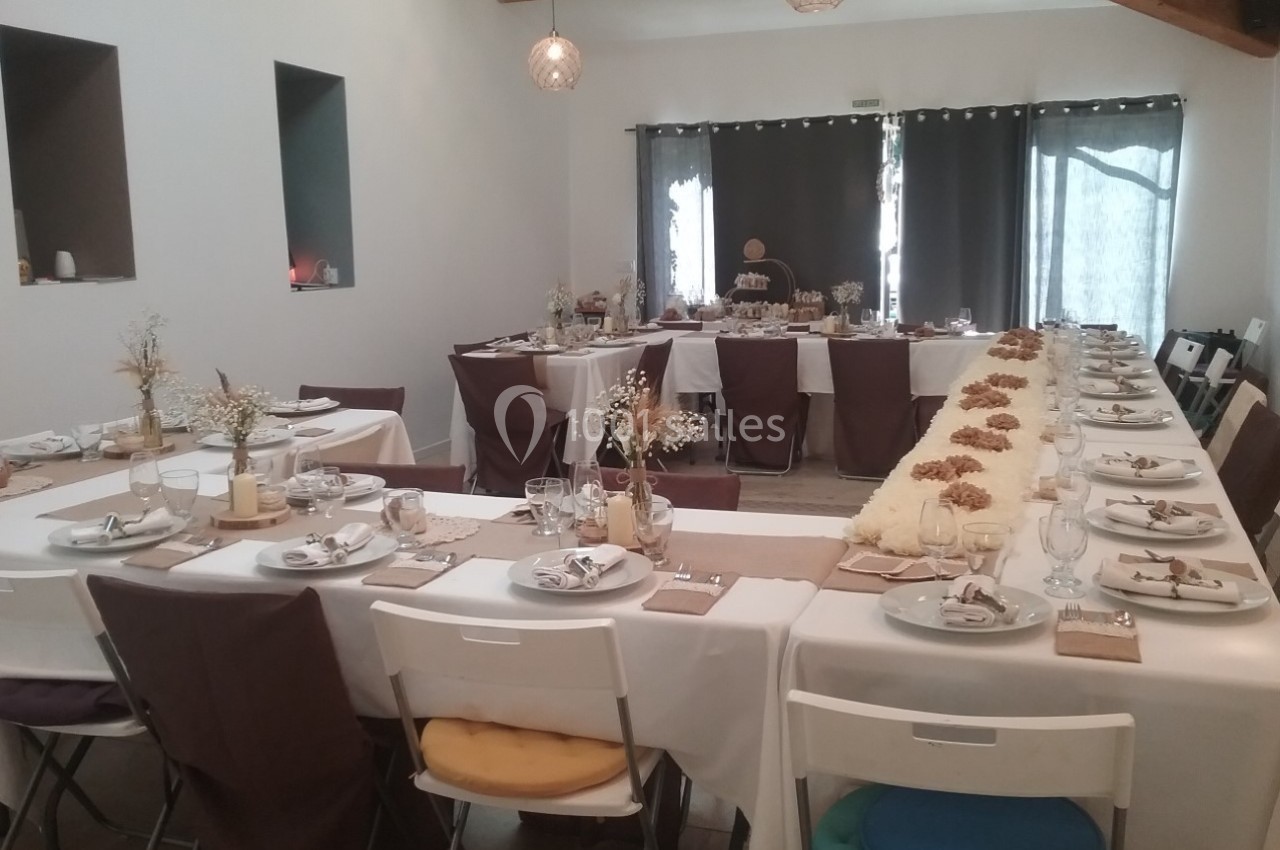 Salle décorée pour un repas festif avec tables en U, nappes blanches, vaisselle élégante et décorations florales.