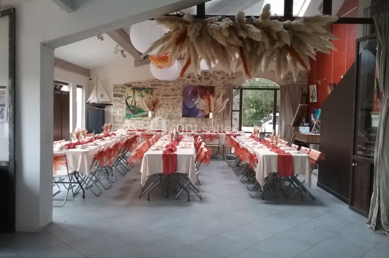 Salle décorée pour un événement avec tables alignées, nappes blanches et chemins de table orange, dans un cadre lumineux.