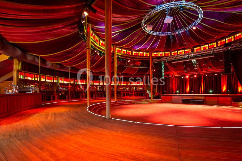 Intérieur d'un chapiteau circulaire avec un sol en bois, éclairage rouge et une scène en arrière-plan.