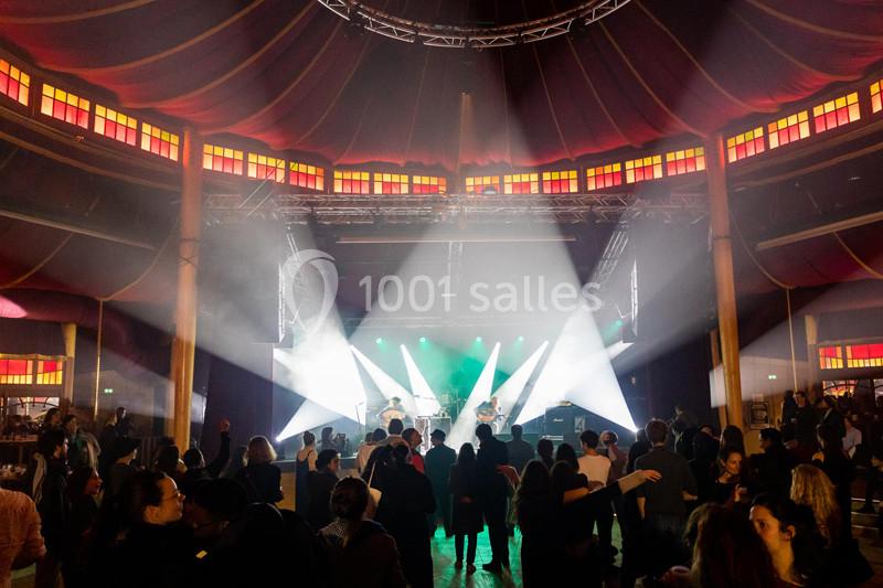 Des spectateurs debout assistent à un concert sous un chapiteau éclairé par des projecteurs colorés.