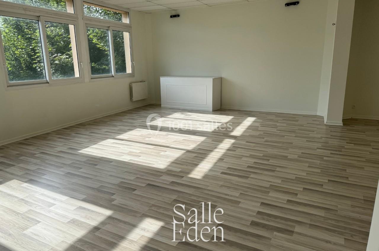 Salle vide avec parquet clair, murs blancs, grandes fenêtres laissant entrer la lumière naturelle et vue sur des arbres.